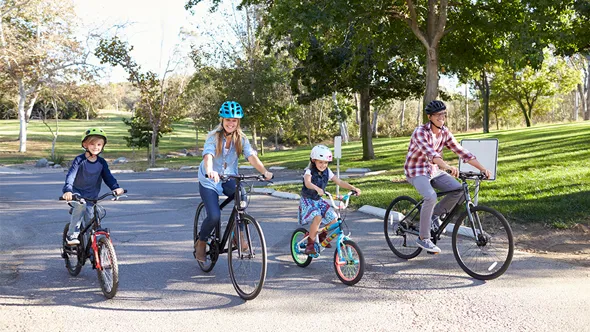 En familj bestående av kvinna, man, pojke, flicka, cyklar tillsammans genom en park på sommaren.
