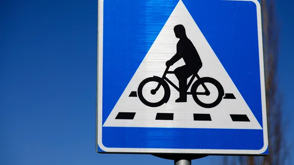 Trafikskylt Cykelöverfart (blå botten, vit liksidig triangel med svart figur som cyklar) mot blå himmel.