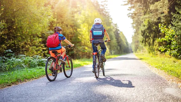 Två skolbarn cyklar på cykelväg med cykelhjälm och ryggsäck.