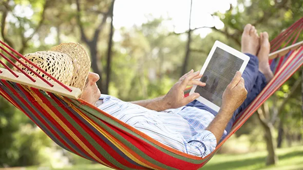 A man in a straw hat lies in a striped hammock in the garden, scrolling on a tablet.